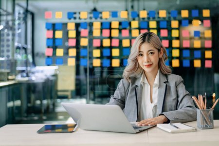 Foto de Joven atractiva Asiática trabajadora de oficina en traje de negocios sonriendo a cámara en oficina moderna - Imagen libre de derechos