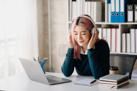 Foto de Smiling asian girl relaxing at home, she is playing music using  laptop, and wearing white headphones - Imagen libre de derechos