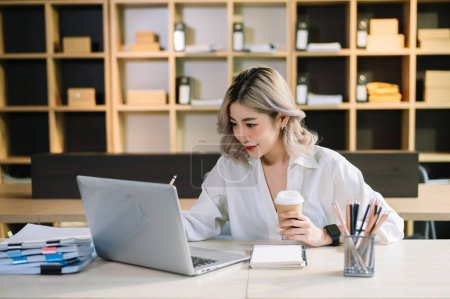 Foto de Confident beautiful Asian businesswoman with laptop  while holding coffee at modern office - Imagen libre de derechos