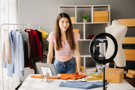 Photo for Fashion blogger concept, Young Asian woman selling clothes on video streaming.Startup small business SME, using smartphone and tablet taking receive and checking in office - Royalty Free Image