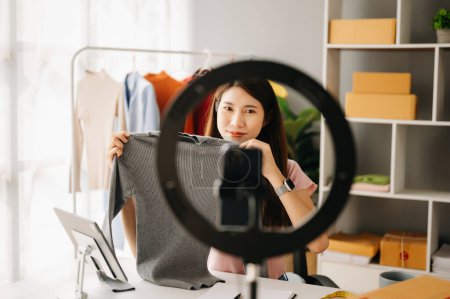 Téléchargez les photos : Concept de blogueur de mode, Jeune femme asiatique vendant des vêtements sur vidéo streaming.Startup PME de petite entreprise, en utilisant smartphone ou tablette prendre recevoir et vérifier au bureau - en image libre de droit