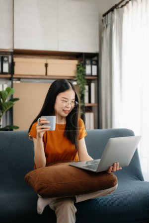 Téléchargez les photos : Asiatique fille assis sur le canapé en utilisant un ordinateur portable dans le salon - en image libre de droit