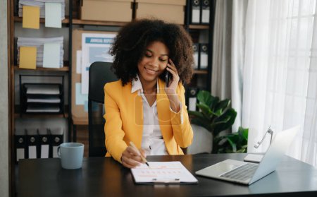 Téléchargez les photos : Femme africaine d'affaires Parler au téléphone et utiliser un ordinateur portable au bureau moderne - en image libre de droit