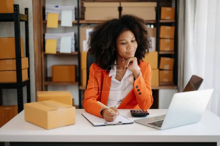 Photo for Startup small business SME, Entrepreneur owner African woman using laptop and checking online purchase shopping order to prepare pack product boxes. at home office - Royalty Free Image