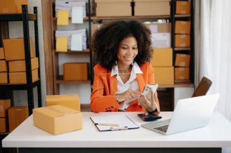 Photo for Startup small business SME, Entrepreneur owner African woman using smartphone and laptop taking receive and checking online purchase shopping order to prepare pack product boxes at home office - Royalty Free Image