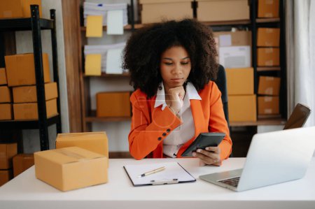 Foto de Startup PYME pequeña empresa, Empresario propietario mujer africana utilizando el ordenador portátil y comprobar la compra en línea orden de compra para preparar cajas de productos de paquete. en la oficina en casa - Imagen libre de derechos