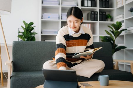 Téléchargez les photos : Femme asiatique confiante avec bloc-notes et tablette PC dans le salon sur le canapé au bureau à la maison - en image libre de droit