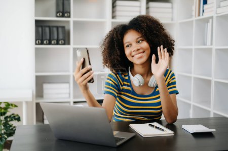Téléchargez les photos : Belle femme africaine utilisant ordinateur portable et téléphone tout en étant assis à son lieu de travail. Concentré au bureau à domicile - en image libre de droit