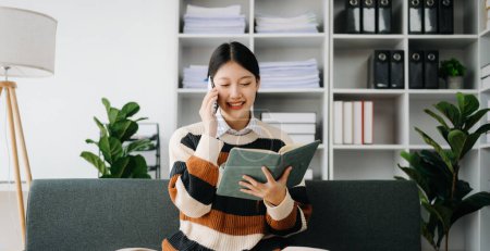 Téléchargez les photos : Femme asiatique confiante avec bloc-notes et téléphone dans le salon sur le canapé au bureau à la maison - en image libre de droit