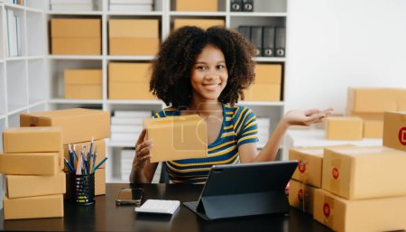 Foto de Startup pyme pequeña empresa, Empresario propietario mujer africana usando portátil tomar recibir y comprobar la compra en línea orden de compra para preparar cajas de productos paquete en la oficina - Imagen libre de derechos