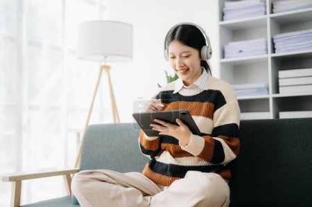 Photo for Attractive happy young Asian student studying  at the sofa, using tablet and headphones having a video chat. - Royalty Free Image