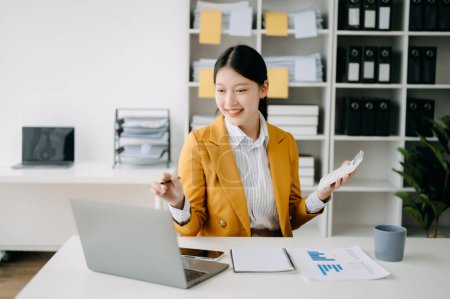 Téléchargez les photos : Jeune femme asiatique attrayante avec ordinateur portable, portable et téléphone travaillant au bureau moderne - en image libre de droit
