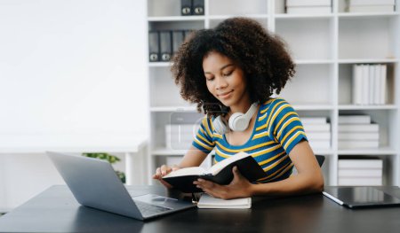 Téléchargez les photos : Étudiante, fille africaine assise au bureau en utilisant un ordinateur portable et un livre - en image libre de droit