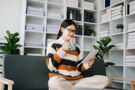 Téléchargez les photos : Femme asiatique confiante avec bloc-notes et café dans le salon sur le canapé au bureau à la maison - en image libre de droit