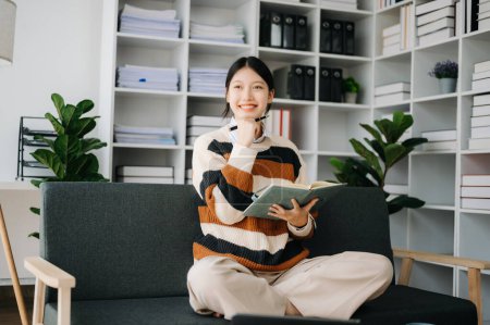 Téléchargez les photos : Femme asiatique confiante tenant un bloc-notes dans le salon sur le canapé au bureau à la maison - en image libre de droit