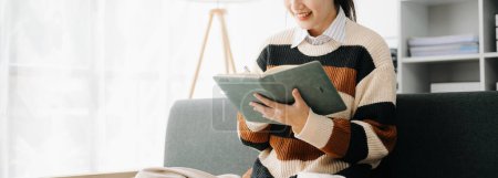 Téléchargez les photos : Jeune femme asiatique note d'écriture pour son idée de travail dans un journal intime dans le salon sur le canapé au bureau à la maison - en image libre de droit