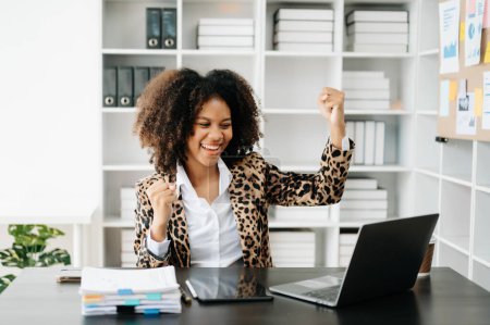 Téléchargez les photos : Femme d'affaires africaine ravie et heureuse et travaillant sur un ordinateur portable au bureau moderne - en image libre de droit