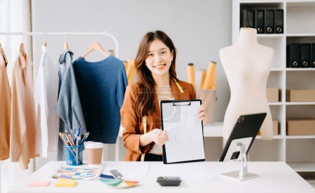 Téléchargez les photos : Asiatique tailleur femme travaillant sur vêtements atelier. ayant un appel vidéo et montrant le dossier avec des statistiques - en image libre de droit