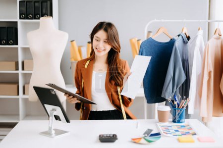 Téléchargez les photos : Jeune tailleur asiatique travaillant sur des vêtements dans un atelier de couture. ayant un appel vide et montrant le dossier avec des statistiques - en image libre de droit