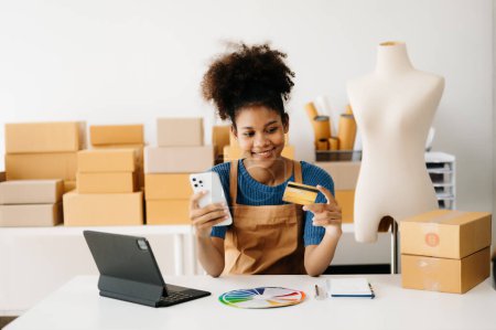 Photo for Entrepreneur owner. African American woman using smartphone and tablet at desk, taking receive and checking online purchase shopping order to preparing pack product boxes - Royalty Free Image