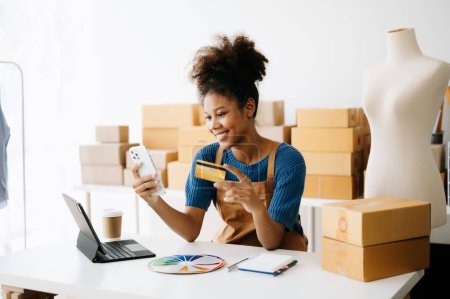 Photo for Happy and smiling African-American woman using smartphone while sitting at desk with laptop, show room with purchase boxes - Royalty Free Image