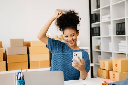 Téléchargez les photos : Femme afro-américaine heureuse et souriante utilisant un smartphone tout en étant assise au bureau avec un ordinateur portable, salle d'exposition avec des boîtes d'achat - en image libre de droit