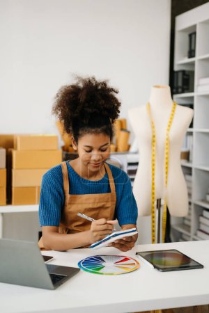 Photo for African-American woman making notes with pen while sitting at desk with laptop. shipping packages boxes on background - Royalty Free Image
