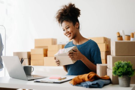 Téléchargez les photos : Femme afro-américaine préparer des boîtes de colis pour la livraison en ligne tout en étant assis au bureau avec ordinateur portable - en image libre de droit