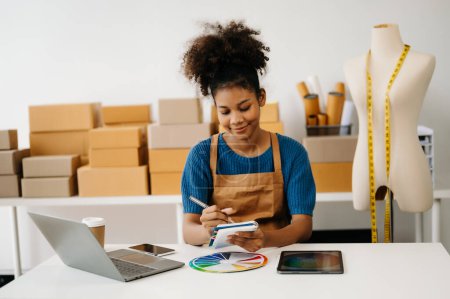 Photo for African-American woman making notes with pen while sitting at desk with laptop. shipping packages boxes on background - Royalty Free Image