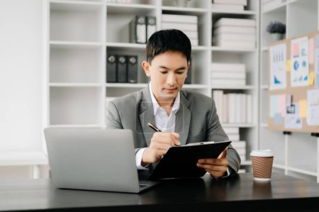 Téléchargez les photos : Jeune homme d'affaires asiatique travaillant au bureau moderne avec ordinateur portable et prenant des notes sur le papier - en image libre de droit