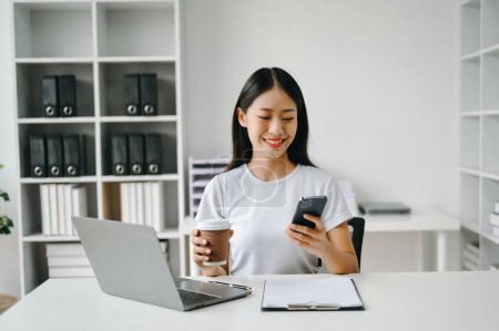 Téléchargez les photos : Asiatique Businesswoman en utilisant le téléphone mobile dans le bureau et tenant tasse de café - en image libre de droit
