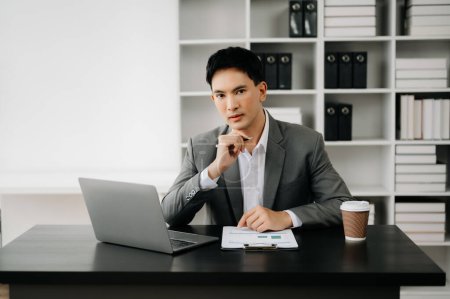 Téléchargez les photos : Jeune homme d'affaires asiatique travaillant au bureau moderne avec ordinateur portable, tablette et prendre des notes sur le papier - en image libre de droit