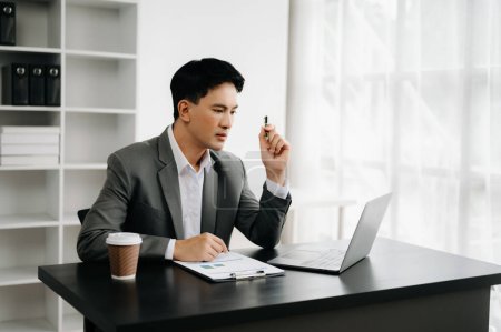 Téléchargez les photos : Jeune homme d'affaires asiatique travaillant au bureau moderne avec ordinateur portable et prenant des notes sur le papier - en image libre de droit