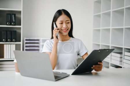 Téléchargez les photos : Asiatique Businesswoman en utilisant le téléphone mobile dans le bureau, souriant et tenant dossier - en image libre de droit