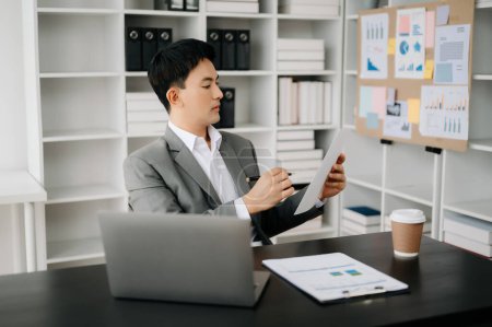 Foto de Joven hombre de negocios asiático que trabaja en la oficina moderna con ordenador portátil, tableta y tomando notas en el papel - Imagen libre de derechos