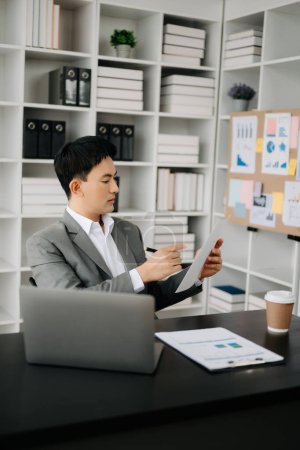 Foto de Joven hombre de negocios asiático que trabaja en la oficina moderna con ordenador portátil, tableta y tomando notas en el papel - Imagen libre de derechos