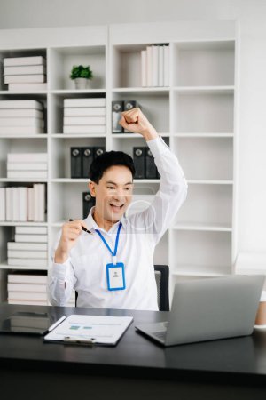 Téléchargez les photos : Heureux homme d'affaires asiatique se sentant excité par son succès professionnel. Jeune homme faisant le geste du gagnant au bureau - en image libre de droit