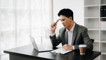 Foto de Joven hombre de negocios asiático trabajando en la oficina moderna con el ordenador portátil y tomando notas en el papel - Imagen libre de derechos