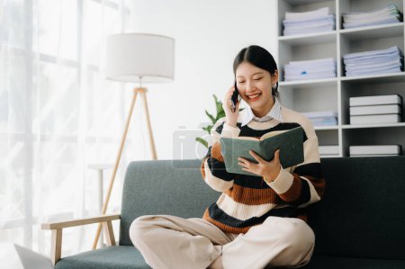 Téléchargez les photos : Confiant asiatique femme lecture livre dans le salon sur le canapé au bureau à domicile - en image libre de droit