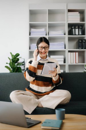 Photo for Attractive happy young Asian student studying at the college library, sitting at the sofa, using smartphone - Royalty Free Image
