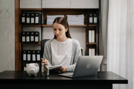 Foto de Mujer financiera de negocios haciendo notas en el escritorio de la oficina con alcancía, monedas de dinero portátil y calculadora - Imagen libre de derechos