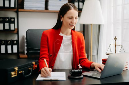 Photo for Caucasian lawyer woman wearing red jacket, working at office desk with laptop and writing notes with pen - Royalty Free Image