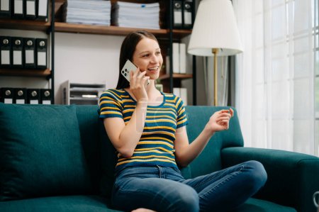 Photo for Smiling Caucasian woman talking on mobile phone while sitting on sofa in Living Room - Royalty Free Image