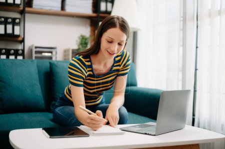 Téléchargez les photos : Freelance femme caucasienne écrit des notes avec stylo dans un carnet en papier, table avec ordinateur portable. travail à domicile - en image libre de droit