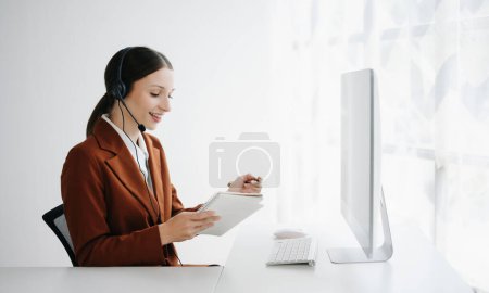 Photo for Call center operator in wireless headset talking with customer and writing notes, woman in headphones with microphone consulting client while working at table with computer monitor - Royalty Free Image
