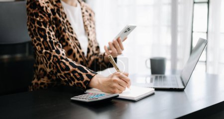 Photo for Cropped image of woman wearing leopard jacket, working at office desk, using mobile phone and writing notes with pen - Royalty Free Image