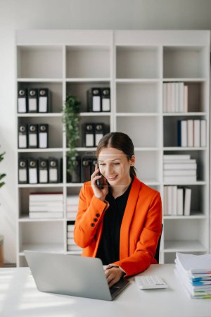 Téléchargez les photos : Business Femme caucasienne Parler au téléphone et utiliser un ordinateur portable avec un sourire tout en étant assis au bureau moderne - en image libre de droit