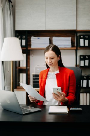 Photo for Caucasian business woman wearing red jacket, working at office desk with laptop and using mobile phone, - Royalty Free Image