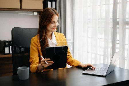 Foto de Experta en negocios segura atractiva sonriente joven asiática sosteniendo tableta digital en el escritorio en la oficina creativa. - Imagen libre de derechos