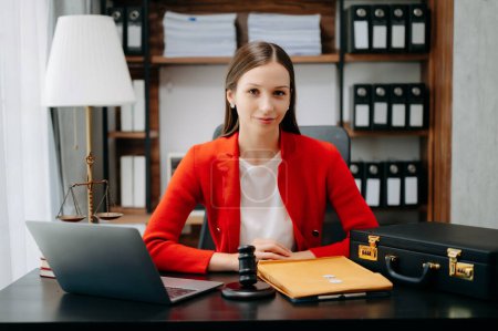 Foto de Mujer abogada caucásica con chaqueta roja y sentada en el escritorio de la oficina con laptop, martillo, estuche y carpeta, - Imagen libre de derechos